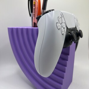 Puede incluir: Un organizador de escritorio curvo de rayas moradas con un mando de PlayStation 5 blanco descansando en la parte superior. El mando está orientado hacia el lado derecho de la imagen.