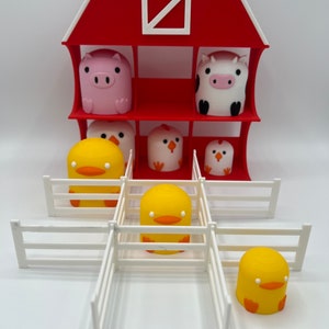 Pode incluir: Um celeiro vermelho com uma janela branca e uma cerca branca. Há quatro figuras de animais dentro do celeiro: um porco, uma vaca, um pintinho e um pintinho. Há três figuras de patos amarelos fora do celeiro.