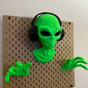 May include: A green alien figurine with headphones on, wearing a green shirt and holding out its hands. The figurine is mounted on a pegboard.