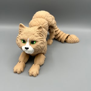Könnte beinhalten: Eine beige Katzenfigur mit grünen Augen und einem weißen Fleck im Gesicht. Die Figur ist in einer hockenden Position mit dem Schwanz hinter sich eingerollt. Der Körper der Katze ist segmentiert, was die Artikulation ermöglicht.