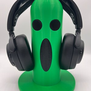 Op de afbeelding: Een groene 3D-geprinte koptelefoonstandaard in de vorm van een gezicht met een grote mond en twee ogen. De standaard houdt een zwarte koptelefoon.