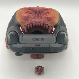 May include: A 3D printed red and orange demon head controller stand with a black Xbox controller resting in its mouth. A red and black dice is in the foreground.