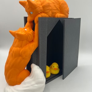 Puede incluir: Una figurita de zorro naranja impresa en 3D con una cola blanca, trepando por un libro negro con una figurita de zorro blanco en la parte superior. Dos patitos de goma amarillos son visibles dentro del libro.
