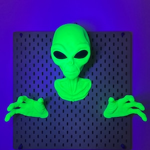 May include: A neon green alien head and two matching hands mounted on a dark gray rectangular pegboard. The alien features large, smooth, almond-shaped black eyes, a small nose, and a defined neck structure, with hands positioned on either side.