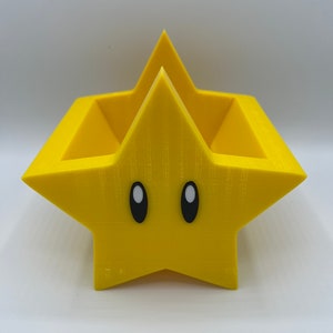 Puede incluir: Un macetero amarillo en forma de estrella con un centro hueco y ojos negros. El macetero está hecho de plástico impreso en 3D.