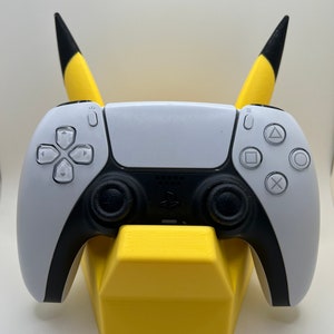May include: A yellow Pikachu-themed stand with black ears holds a white and black PlayStation 5 controller.