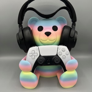 Pode incluir: Uma figura de urso colorido com um design de gradiente de arco-íris, usando fones de ouvido pretos e segurando um controle de jogo branco. O design do urso inclui cores pastel como rosa, azul e amarelo.