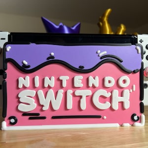 May include: A Nintendo Switch console with a custom pink and purple faceplate. The faceplate features the words "Nintendo Switch" in white, outlined in black. The console has custom thumb grips, one with a paw print and the other with a Kirby design.