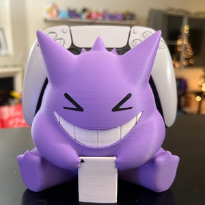 May include: A purple and white 3D printed Ghost Pokemon figure holding a white controller stand. The figure has a wide grin and is sitting on a black surface.