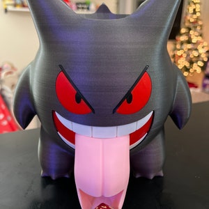K&ouml;nnte beinhalten: Eine schwarz-graue 3D-gedruckte Gengar-Figur mit roten Augen und einer rosa Zunge. Die Figur ist so konzipiert, dass sie einen W&uuml;rfel h&auml;lt, der auf dem Bild als roter und goldener W6 mit der Zahl 6 nach oben dargestellt ist.
