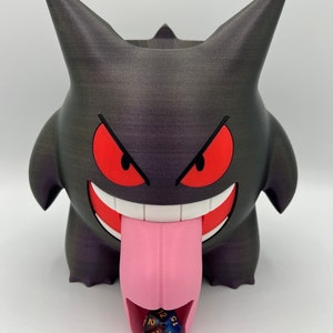 K&ouml;nnte beinhalten: Ein schwarz-violetter 3D-gedruckter W&uuml;rfeltower in Form eines Gengar-Pok&eacute;mon mit einer rosa Zunge und einem roten Mund. Ein blauer und goldener W&uuml;rfel rollt aus der Zunge.
