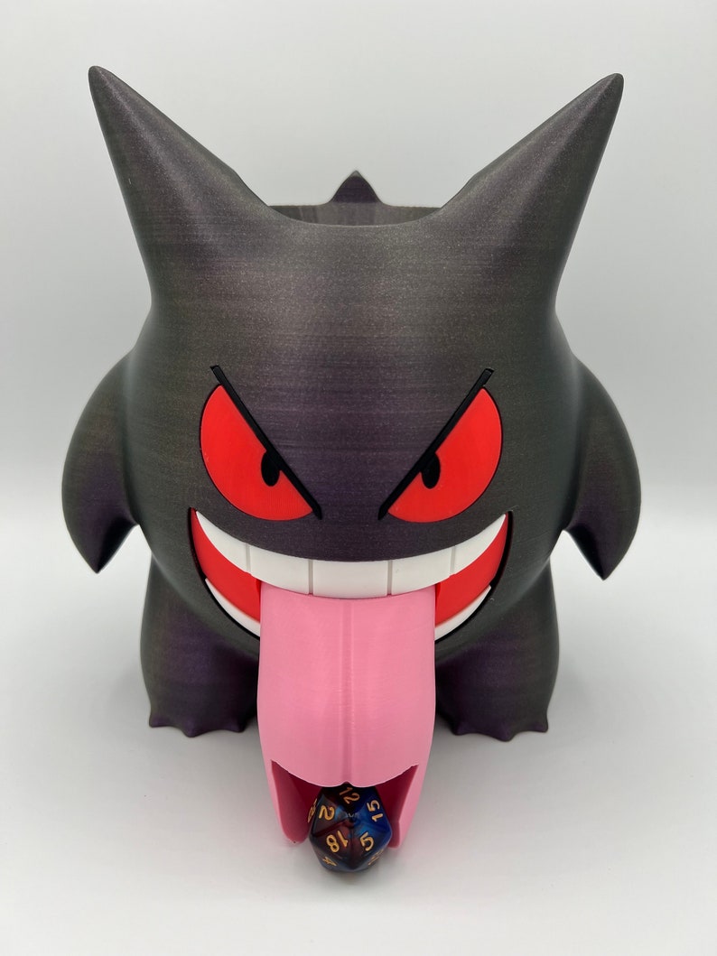 K&ouml;nnte beinhalten: Ein schwarz-violetter 3D-gedruckter W&uuml;rfeltower in Form eines Gengar-Pok&eacute;mon mit einer rosa Zunge und einem roten Mund. Ein blauer und goldener W&uuml;rfel rollt aus der Zunge.