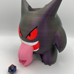 K&ouml;nnte beinhalten: Ein 3D-gedruckter schwarzer und lilafarbener Gengar-W&uuml;rfelhalter mit einer rosa Zunge. Der Halter ist so konzipiert, dass er W&uuml;rfel auff&auml;ngt, wenn sie geworfen werden. Ein blauer und goldener W&uuml;rfel befindet sich vor dem Halter.