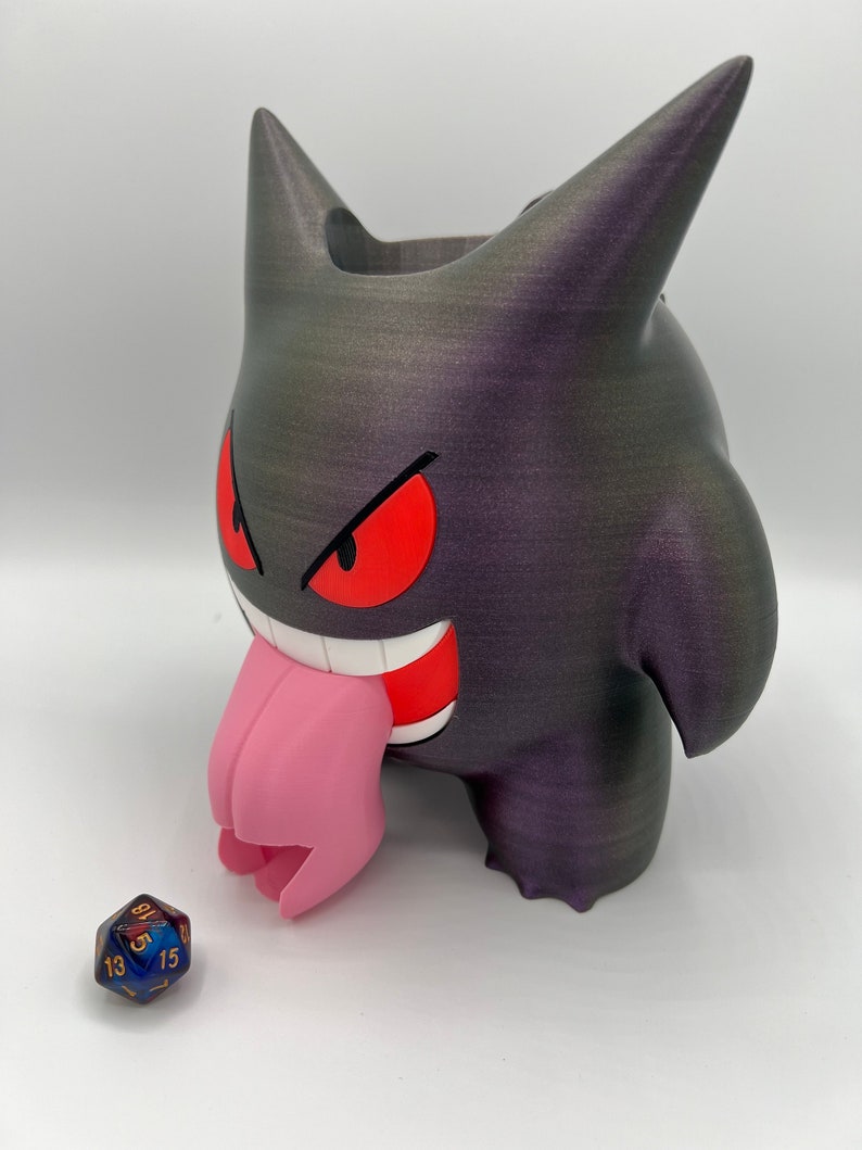 K&ouml;nnte beinhalten: Ein 3D-gedruckter schwarzer und lilafarbener Gengar-W&uuml;rfelhalter mit einer rosa Zunge. Der Halter ist so konzipiert, dass er W&uuml;rfel auff&auml;ngt, wenn sie geworfen werden. Ein blauer und goldener W&uuml;rfel befindet sich vor dem Halter.