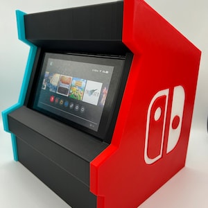 May include: A 3D printed Nintendo Switch stand in the shape of an arcade cabinet. The stand is black, red, and blue. The Switch is in the stand and the screen is showing the home screen with several game icons. The stand has the Nintendo Switch logo on the side.