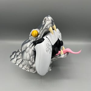 Peut inclure: Un support de manette en forme de tête de serpent imprimé en 3D argenté avec une langue rose et des yeux jaunes. Deux manettes de jeu vidéo blanches sont tenues dans la gueule du serpent.