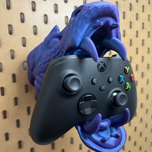 May include: A 3D printed purple dragon head wall mount for a black Xbox controller.