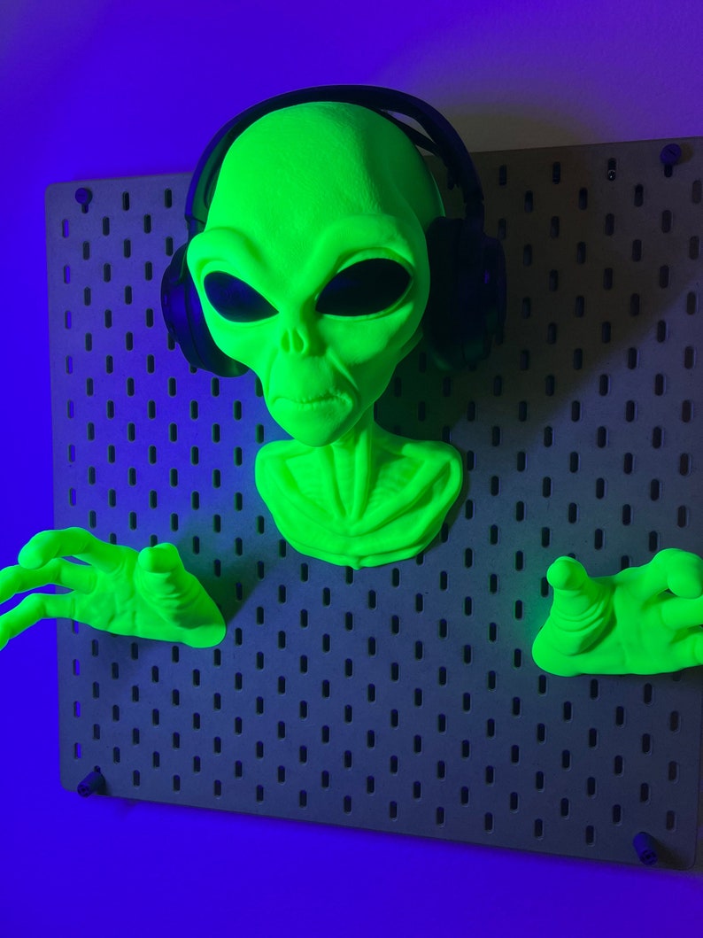 May include: A bright neon green alien head and two hands mounted on a dark gray pegboard. The alien figure wears black over-ear headphones. The head features large, dark, almond-shaped eyes and a small nose, while the hands are outstretched.