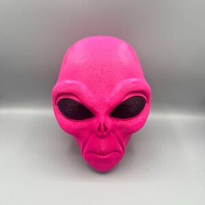 May include: A bright neon pink alien head sculpture featuring a smooth, textured surface with large, dark, oval-shaped eyes. The figure displays a distinct brow ridge, a small nose area with two nostrils, and a thin, straight mouth line.