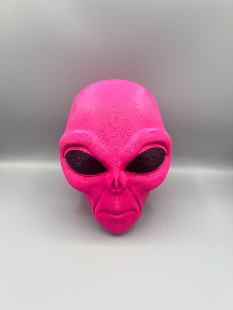 May include: A bright neon pink alien head sculpture featuring a smooth, textured surface with large, dark, oval-shaped eyes. The figure displays a distinct brow ridge, a small nose area with two nostrils, and a thin, straight mouth line.