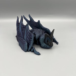 May include: A detailed, dark blue bat figurine with textured wings and body. The bat is lying down, with its wings spread slightly. The figurine has a matte finish and is set against a plain, light gray background.