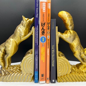 Puede incluir: Un par de sujetalibros de zorro dorados sosteniendo una pila de libros. Los sujetalibros están hechos de un material texturizado y tienen un diseño de zorro realista. Los libros incluyen un libro de texto de idioma japonés, un libro de trabajo y una novela.