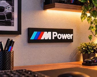 May include: A rectangular wall sign featuring the M Power logo in white text on a black background, accompanied by the iconic blue, dark blue, and red slanted stripes. The sign is mounted on a wall above a wooden desk surface in a workspace.