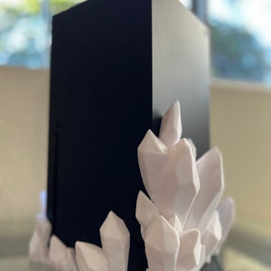 Puede incluir: Un dispositivo electrónico rectangular negro con una base decorativa blanca en forma de cristal. Los cristales están agrupados alrededor de la parte inferior del dispositivo, añadiendo una estética única. El dispositivo está sobre una superficie reflectante.