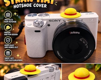 May include: A yellow and red straw hat hotshoe cover for a camera. The cover is shaped like a miniature straw hat with a red band. It is shown attached to the top of a white Sony camera and also displayed separately as a standalone object.