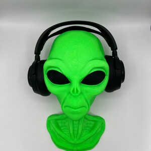 May include: A bright neon green alien head bust featuring large, dark, almond-shaped eyes and a defined neck structure. The alien head is wearing a pair of black over-ear headphones that rest securely on the sides of the head, covering the ear area.