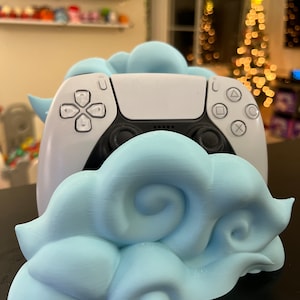 Może przedstawiać: Biały kontroler PlayStation 5 umieszczony jest w stojaku w kształcie chmury, wydrukowanym w 3D w jasnoniebieskim kolorze.