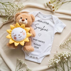 May include: A white baby onesie with the text "OUR LITTLE Sunshine" and "BABY MATEO AUGUST 2026". A brown teddy bear and a yellow sun-shaped plush toy are next to the onesie. White flowers are scattered around the items.