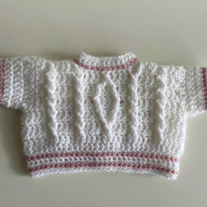 May include: A white, short-sleeved, crocheted sweater with a cable knit design. The sweater has pink trim on the sleeves, neckline, and hem. The handmade garment is likely for a baby or small child.