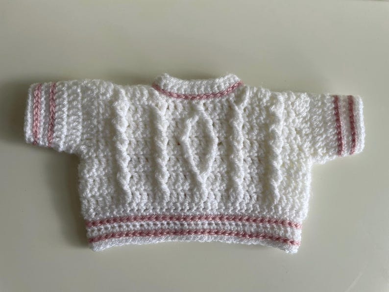 May include: A white, short-sleeved, crocheted sweater with a cable knit design. The sweater has pink trim on the sleeves, neckline, and hem. The handmade garment is likely for a baby or small child.