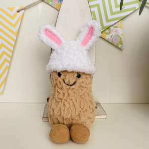 May include: A plush peanut-shaped toy wearing a white bunny hat with pink inner ears. The toy has a smiling face and corduroy feet. Decorative chevron and floral banners are in the background.