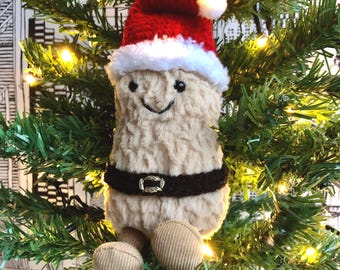 Puede incluir: Adorno navideño de peluche con forma de cacahuete sonriente, con gorro de Papá Noel rojo con ribete blanco y cinturón negro. Tiene pies de pana y está colgado de un árbol de Navidad con luces.