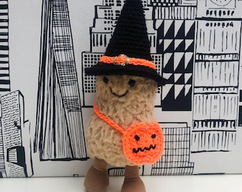 Puede incluir: Un peluche con forma de cacahuete que lleva un sombrero de bruja de ganchillo negro con una banda naranja y una pequeña hebilla dorada. El juguete tiene un cuerpo texturizado de color tostado, pies de tela marrón y un bolso de calabaza naranja.