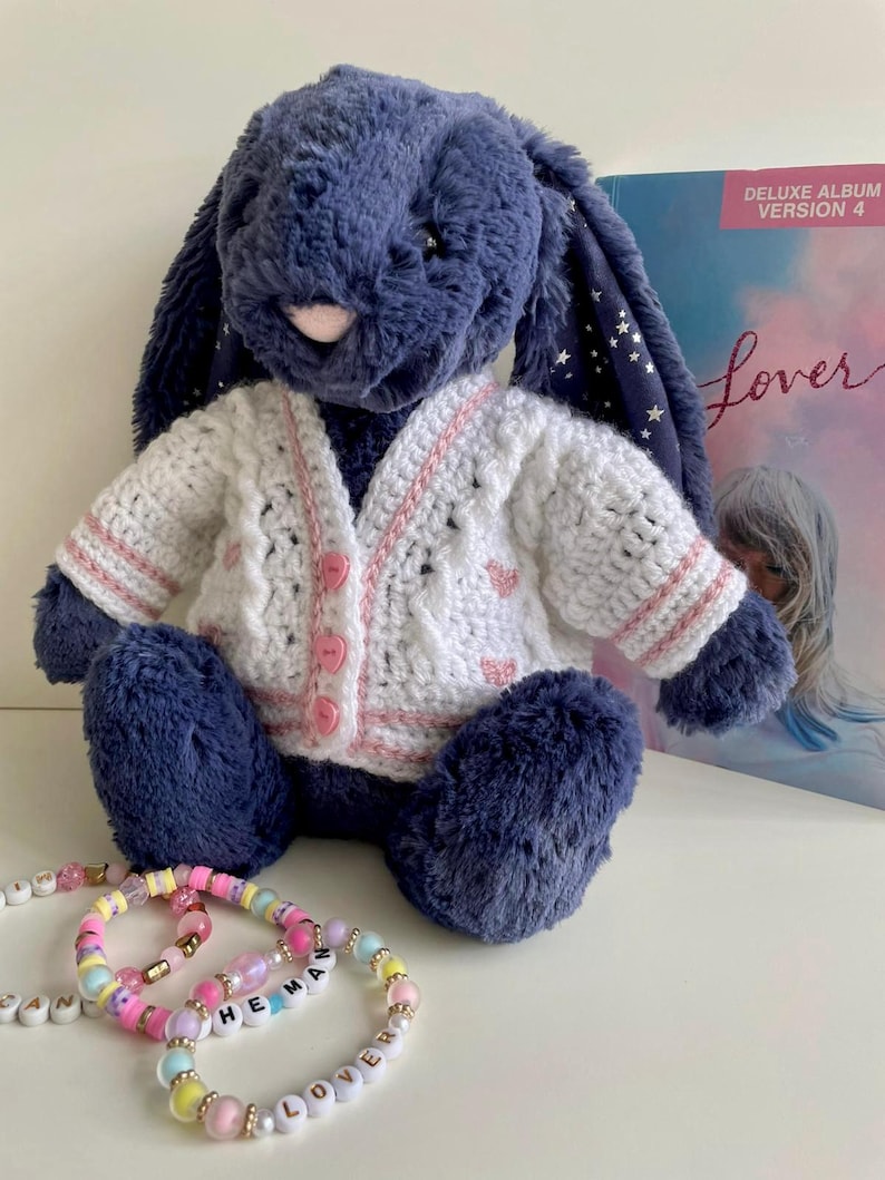 May include: A plush navy blue bunny wearing a white and pink crocheted cardigan. The bunny is sitting with a few colorful beaded bracelets in front of it. A "Lover" album cover is in the background.
