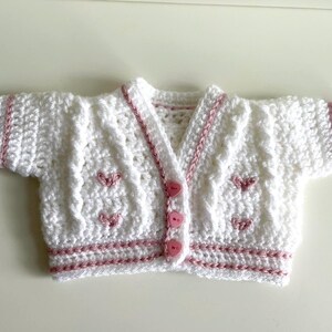 May include: A white, crocheted baby cardigan with pink trim and heart-shaped accents. The short-sleeved sweater has a V-neck and is fastened with three pink buttons. The edges and sleeves are trimmed with pink yarn.