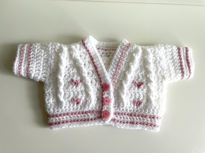 May include: A white, crocheted baby cardigan with pink trim and heart-shaped accents. The short-sleeved sweater has a V-neck and is fastened with three pink buttons. The edges and sleeves are trimmed with pink yarn.