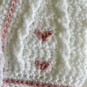 May include: Close-up of a white crocheted item with pink accents. The item features a textured stitch pattern and is adorned with small, pink crocheted hearts. Pink yarn is used for decorative stripes and edging.