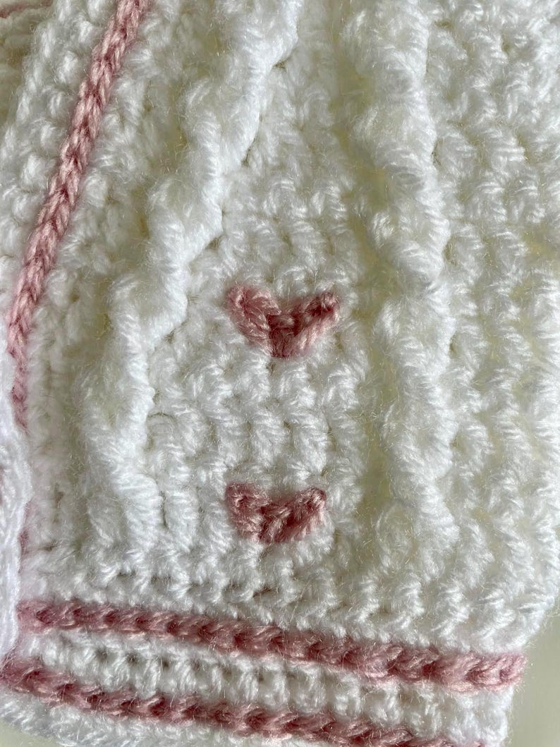 May include: Close-up of a white crocheted item with pink accents. The item features a textured stitch pattern and is adorned with small, pink crocheted hearts. Pink yarn is used for decorative stripes and edging.