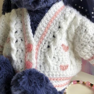 May include: A plush toy wearing a handmade white crochet cardigan with pink trim and heart accents. The cardigan has pink heart-shaped buttons. The toy has blue fur and a bracelet with letter beads.