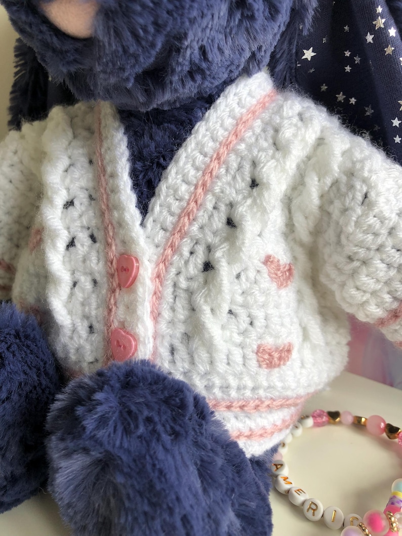 May include: A plush toy wearing a handmade white crochet cardigan with pink trim and heart accents. The cardigan has pink heart-shaped buttons. The toy has blue fur and a bracelet with letter beads.