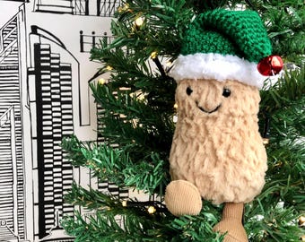 Puede incluir: Un adorno de peluche con forma de cacahuete beige con una cara sonriente bordada, que lleva un gorro de ganchillo verde con un borde de piel sintética blanca y un pequeño cascabel rojo. Dos pies de pana beige cuelgan de la parte inferior del cuerpo.