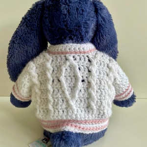 May include: A plush blue bunny wearing a white cable-knit sweater with pink stripes. The bunny's long ears are visible, and a tag is attached to its side. The sweater has a textured design.