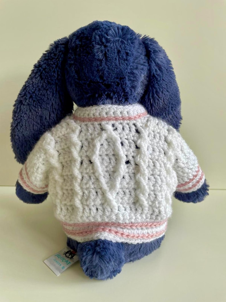 May include: A plush blue bunny wearing a white cable-knit sweater with pink stripes. The bunny's long ears are visible, and a tag is attached to its side. The sweater has a textured design.