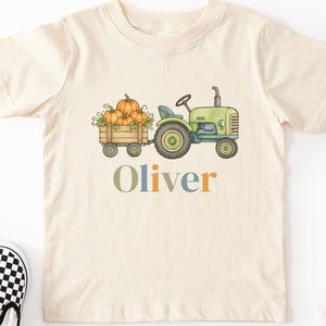 May include: A cream-colored short-sleeve t-shirt featuring a graphic of a green tractor pulling a wooden crate filled with orange pumpkins. The name Oliver is printed below the tractor in multicolored letters. The shirt has a crew neckline.