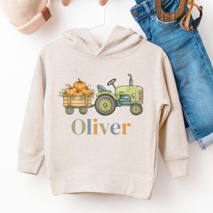 May include: A beige hooded sweatshirt featuring a graphic of a green tractor pulling a wooden cart filled with orange pumpkins. The name Oliver is printed below the graphic in multicolored letters. A beige knit beanie and blue jeans are nearby.