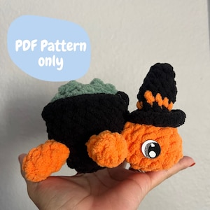 Puede incluir: Una tortuga bruja de Halloween de crochet con cuerpo naranja, caparazón negro y sombrero verde. La tortuga tiene grandes ojos negros con pupilas blancas.